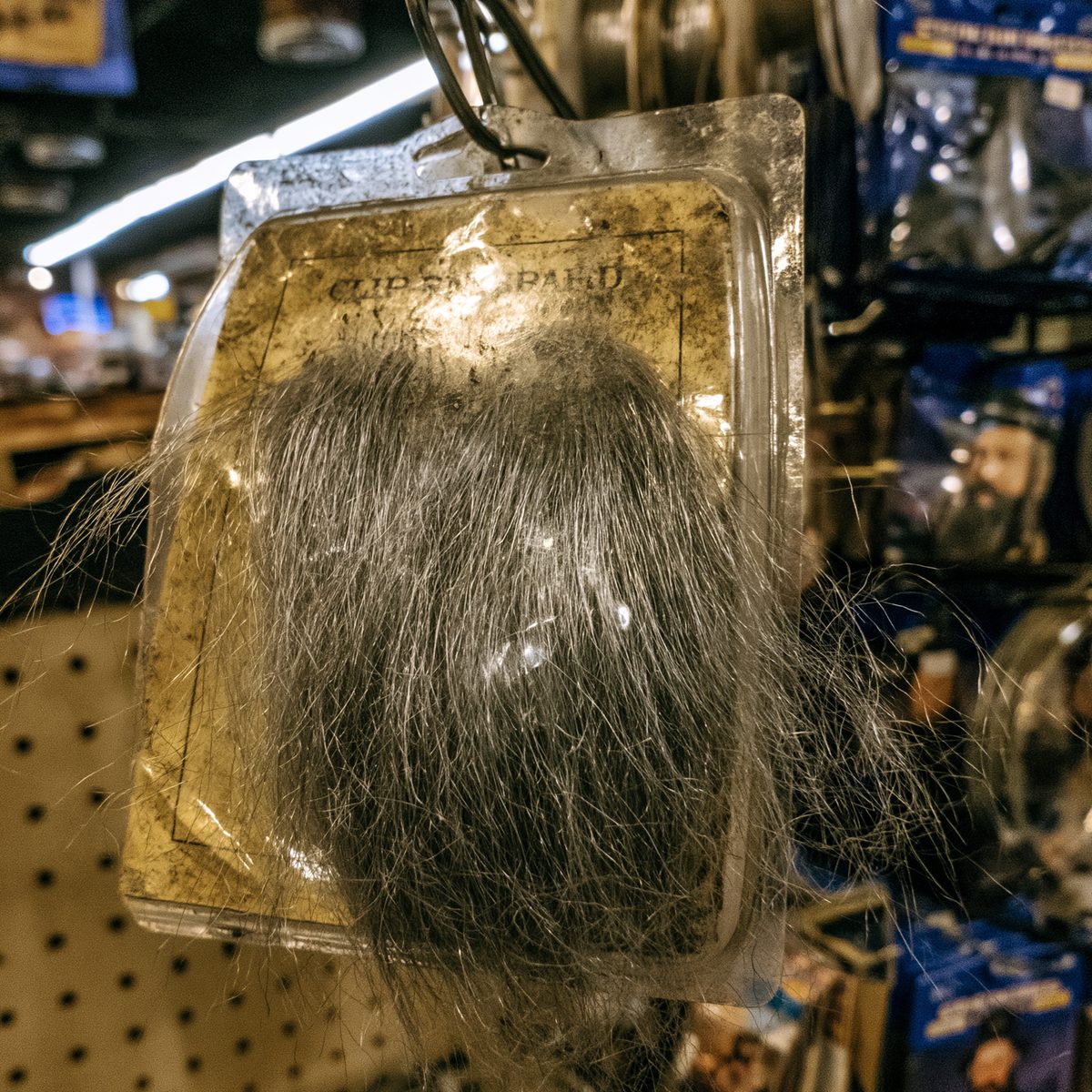 A clip-on polyester beard in a dusty plastic clamshell hanging on a pegboard hook